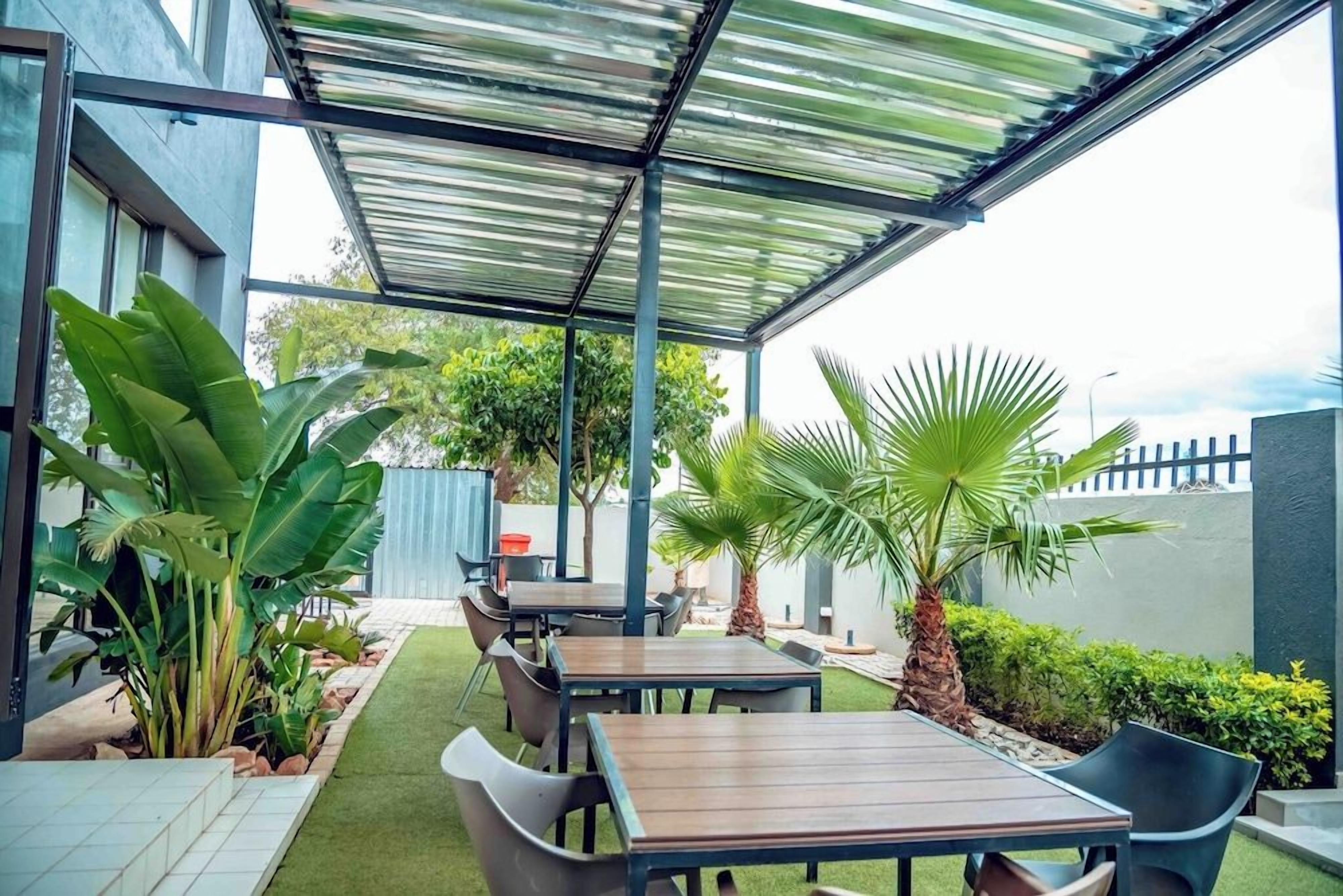 An outdoor patio area with tables and chairs, surrounded by greenery and a covered seating area.