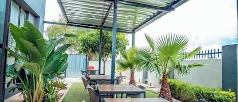 Un patio al aire libre con mesas y sillas, rodeado de follaje y una zona de estar cubierta.