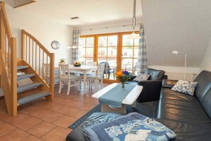 A living room with a staircase, a dining area with a table and chairs, and a sofa with cushions. - 2.5/T3 holiday home park “Freesenbruch” (Zingst (Ostseeheilbad))