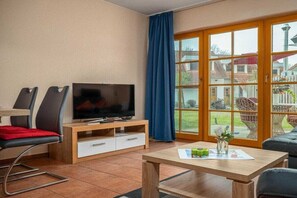A living room with a TV, a dining area with chairs, a table with a vase, and a sliding glass door to the outside. - 1.2/T4 holiday home park “Freesenbruch” (Zingst (Ostseeheilbad))