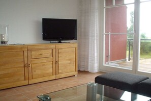 A room with a wooden cabinet, a flat-screen TV, a glass table, and a view through a large window. - Mühlenstraße 19 Apartment 4 (Seeheilbad Zingst)