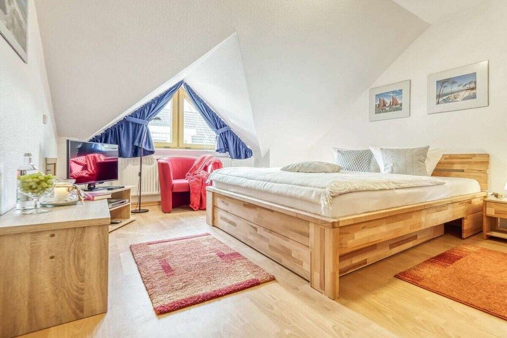 Un dormitorio con cama de madera, un escritorio con silla, una silla roja, una ventana con cortinas azules y una obra de arte en la pared.