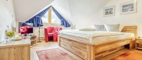 Un dormitorio con cama de madera, un escritorio con silla, una silla roja, una ventana con cortinas azules y una obra de arte en la pared.