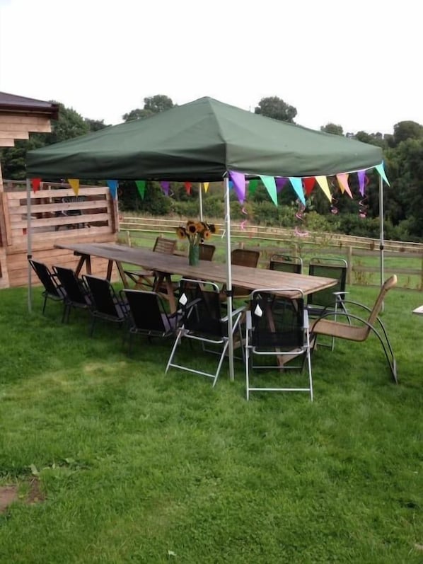 An outdoor dining area with a table and chairs under a green canopy, surrounded by a wooden fence and greenery.