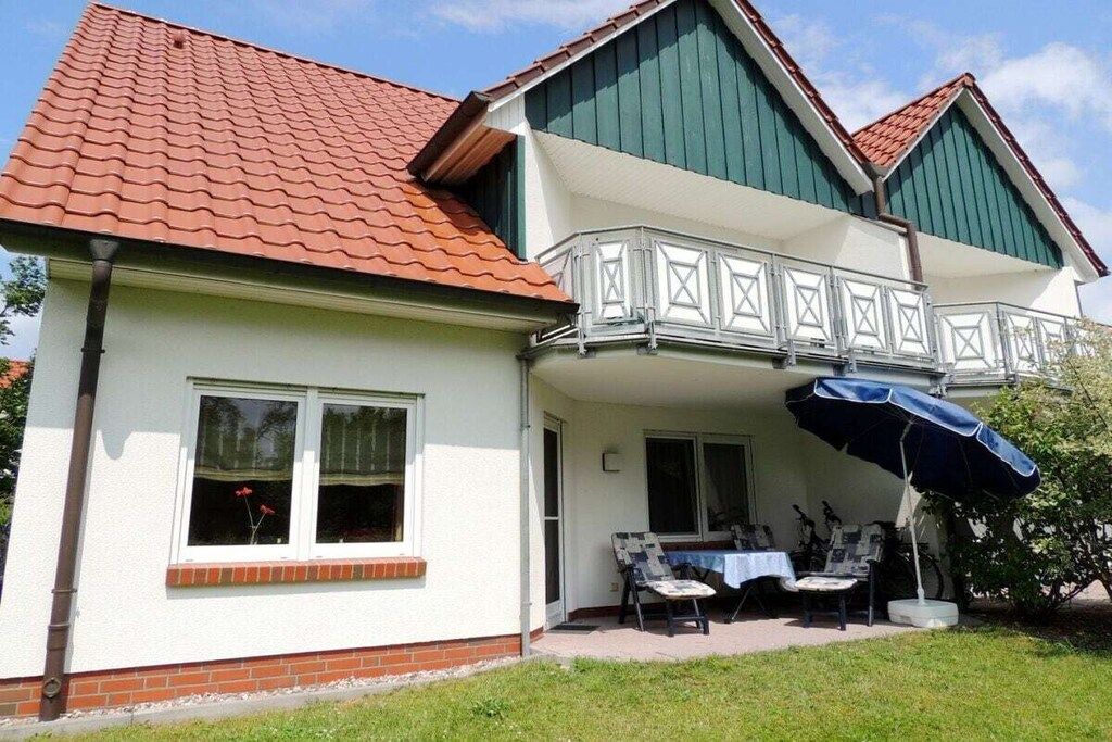 A two-story house with a balcony, a patio with a table and chairs, and a blue umbrella.