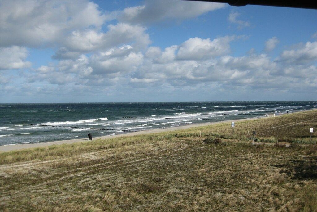 Un paysage côtier avec un premier plan herbeux, une plage de sable et l’océan sous un ciel partiellement nuageux.