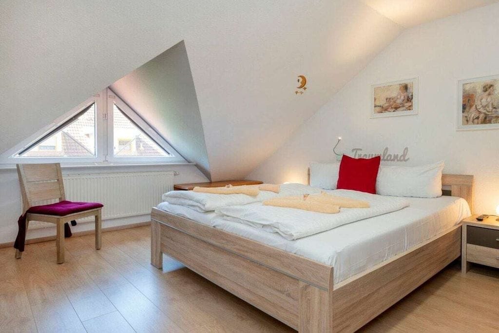 Un dormitorio con techo inclinado, una cama con ropa de cama blanca, un cojín rojo, una silla con un cojín morado y una ventana con vistas a edificios.