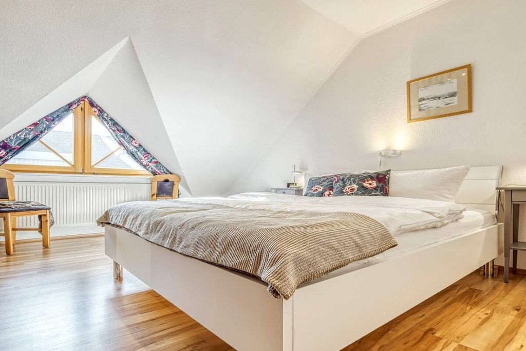 A bedroom with a sloped ceiling, a bed with a patterned throw, a bedside table with a lamp, and a window with a floral curtain.