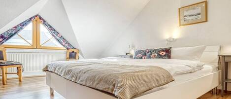A bedroom with a sloped ceiling, a bed with a patterned throw, a bedside table with a lamp, and a window with a floral curtain.