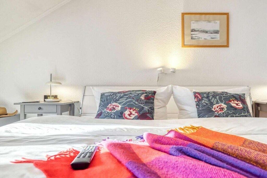 A hotel room with a bed, pillows, a blanket, a remote control, a nightstand, a lamp, and a framed picture on the wall.