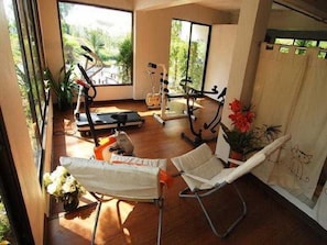 A room with a wooden floor, exercise equipment, a white chair, and a window with a view of greenery.