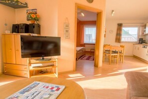 Ein Wohnzimmer mit Flachbildfernseher, Holzmöbeln, einer Essecke mit Stühlen und einer Küche im Hintergrund.