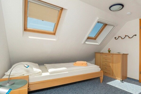 Ein kleines, ordentliches Schlafzimmer mit einem Einzelbett, einem Nachttisch, einer Lampe, einer Kommode und zwei Fenstern.