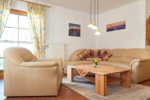Un salon comprenant un canapé, un fauteuil et une table basse. On y trouve deux lampes fixées au mur, un radiateur et deux tableaux encadrés.