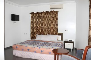 A hotel room with a bed, a TV, a bedside table, a chair, and an air conditioning unit. - Phisbond Hotels (Uyo)