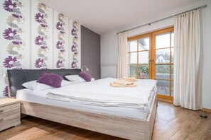 A bedroom with a large bed, a bedside table, a window with curtains, and a view of greenery outside. - 2.1/T2 holiday home park “Freesenbruch” (Zingst (Ostseeheilbad))