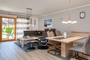 Uma sala de estar moderna com sofá, mesa de centro, mesa de jantar e cadeiras. Há uma janela com cortinas, um lustre e um relógio na parede.