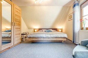 A bedroom with a sloped ceiling, a bed with a wooden headboard, bedside lamps, a chair, and a large mirror. - 2.6/T3 holiday home park “Freesenbruch” (Zingst (Ostseeheilbad))