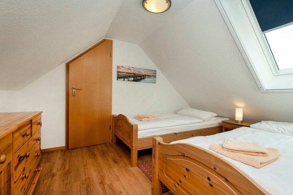Un dormitorio bajo en áticos, de forma triangular, con dos camas de madera, un aparador de madera y una puerta que da a otra habitación.