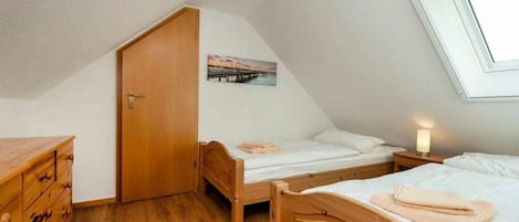 Un dormitorio bajo en áticos, de forma triangular, con dos camas de madera, un aparador de madera y una puerta que da a otra habitación.