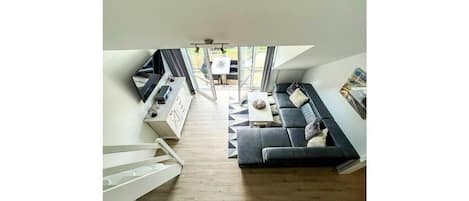Una habitación moderna estilo loft, con suelo de madera, un sofá negro, una mesa de centro blanca y un área de comedor con mesa y sillas.