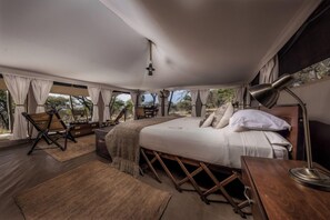 A spacious bedroom with a large bed, a desk, and a chair, featuring a view of the outdoors. - Serengeti Pioneer Camp (Bariadi)