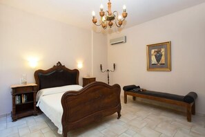 A bedroom with a wooden bed, bedside tables, a bench, a chandelier, and a wall-mounted air conditioner.