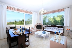 A room with a dining table, chairs, a sofa, a fireplace, and large windows with a view of greenery and mountains.