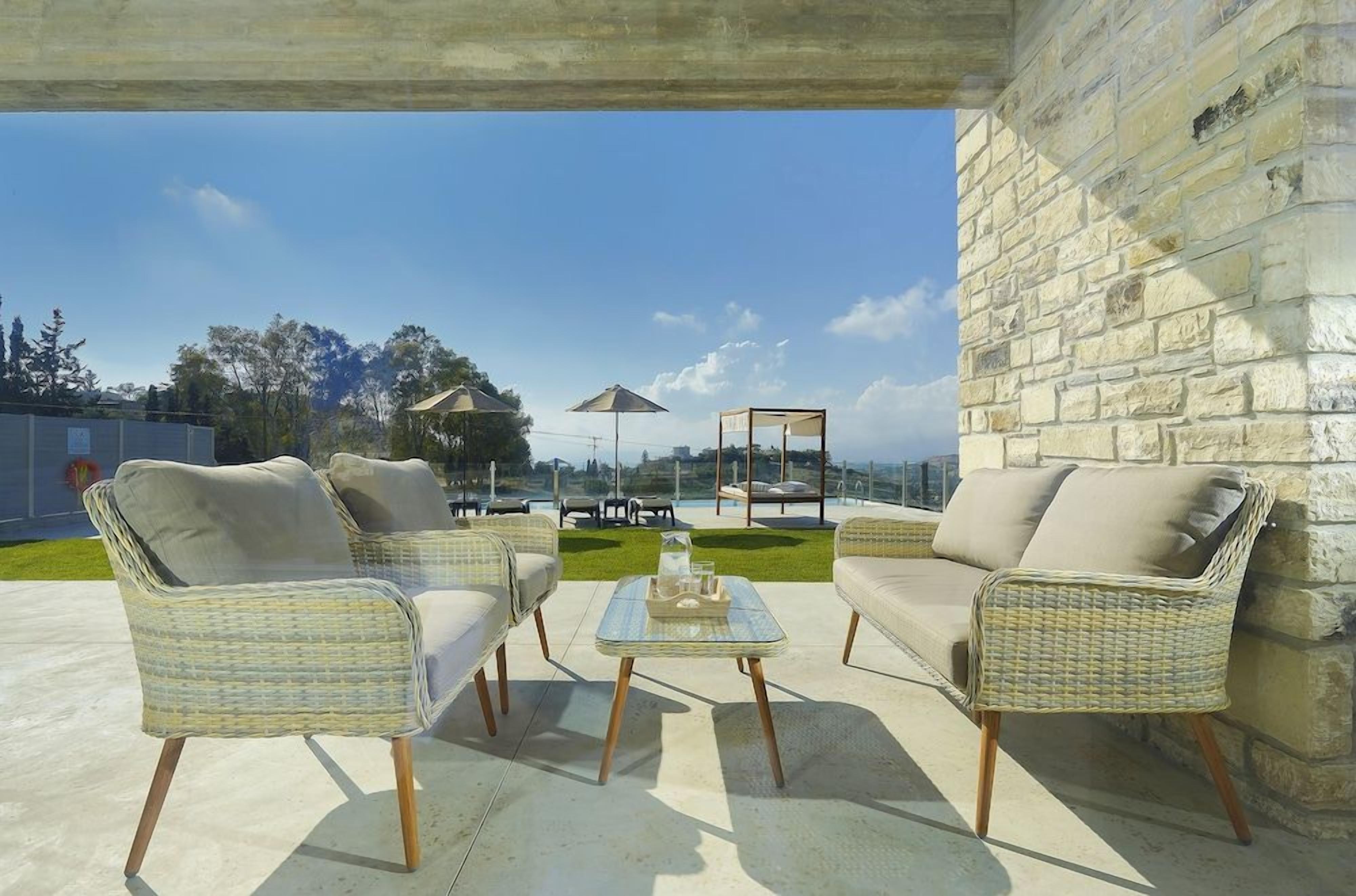 An outdoor seating area with wicker furniture and a glass table, overlooking a pool and greenery.