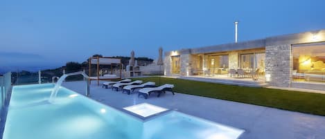 A modern outdoor pool area with lounge chairs, a pergola, and a stone building with outdoor seating.