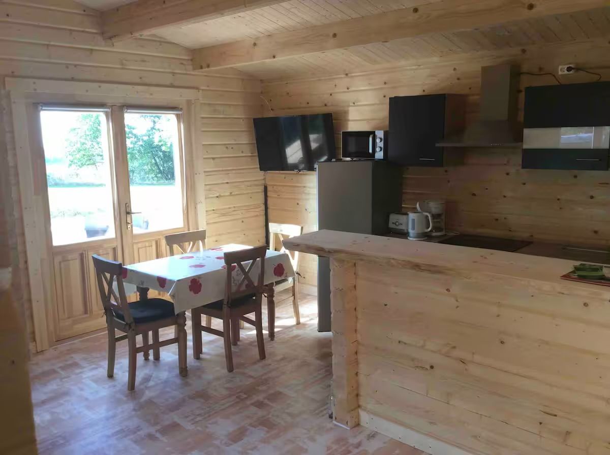 Een houten blokhut interieur met een eettafel, stoelen, een keuken met inbouwapparatuur en een groot raam.