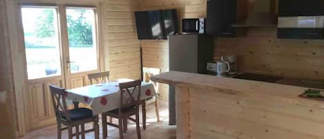Interior de una cabaña de madera con mesa de comedor, sillas, una zona de cocina con electrodomésticos y un ventanal grande.