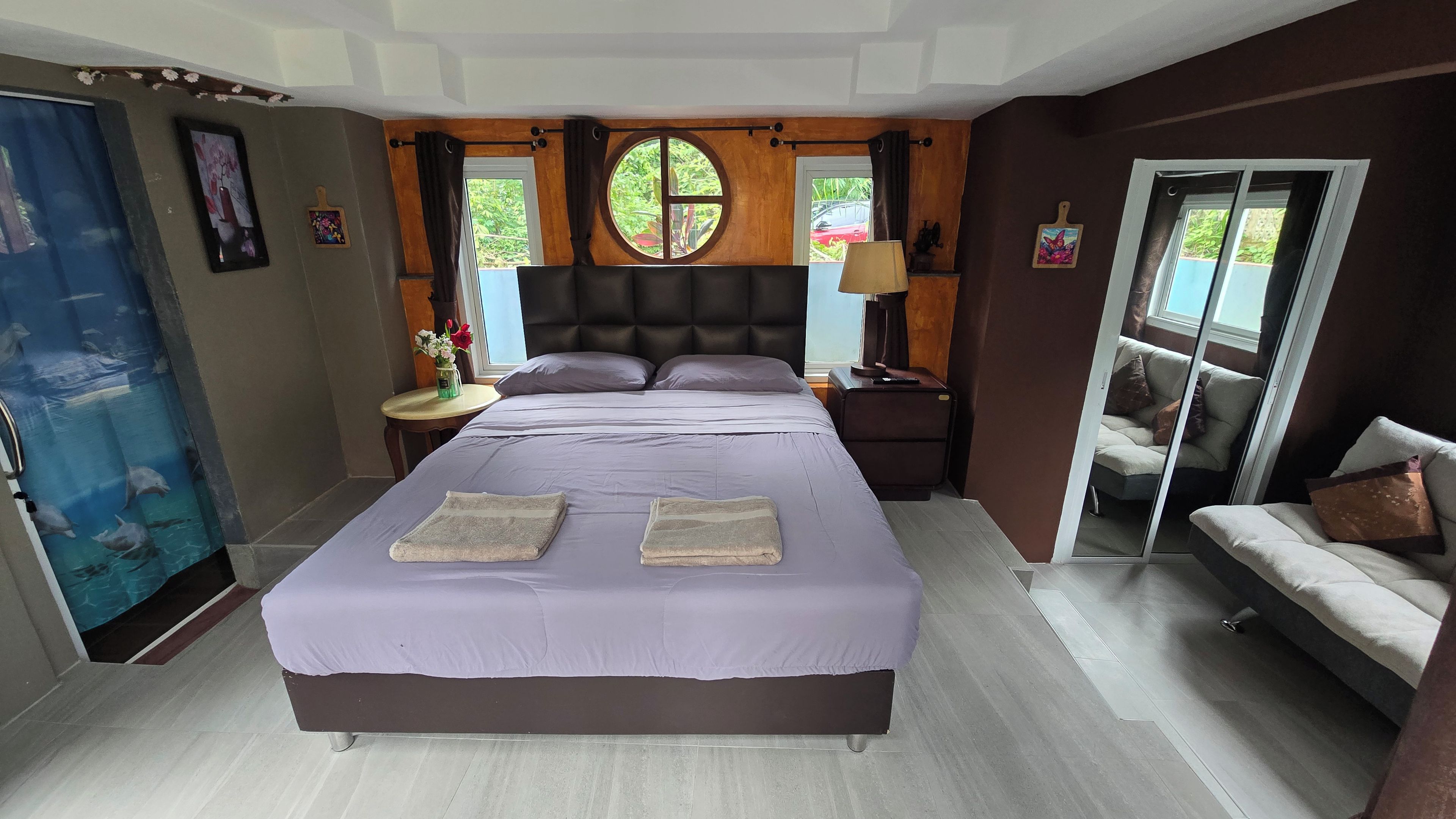 A bedroom with a large bed, a nightstand, a lamp, a sofa, and a window with a view of greenery.