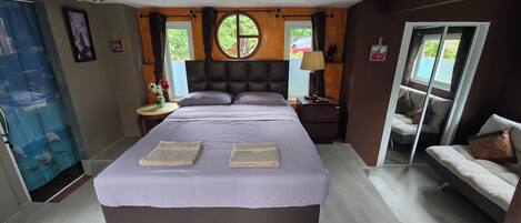A bedroom with a large bed, a nightstand, a lamp, a sofa, and a window with a view of greenery.