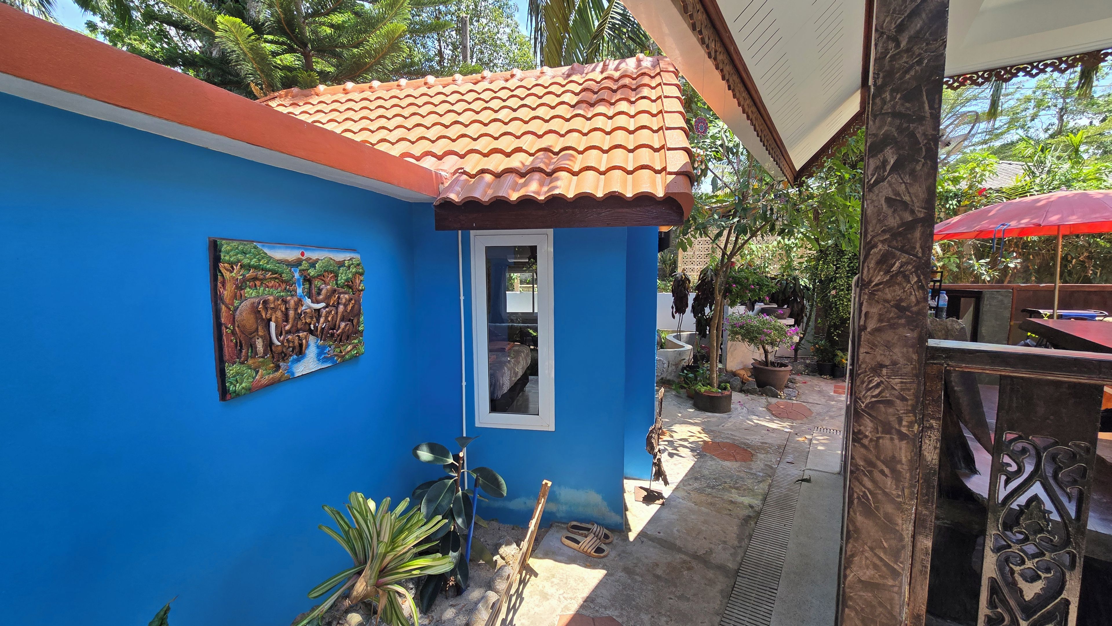 A blue building with a tiled roof, a window, and a door. There is a painting on the wall and a red umbrella in the background.