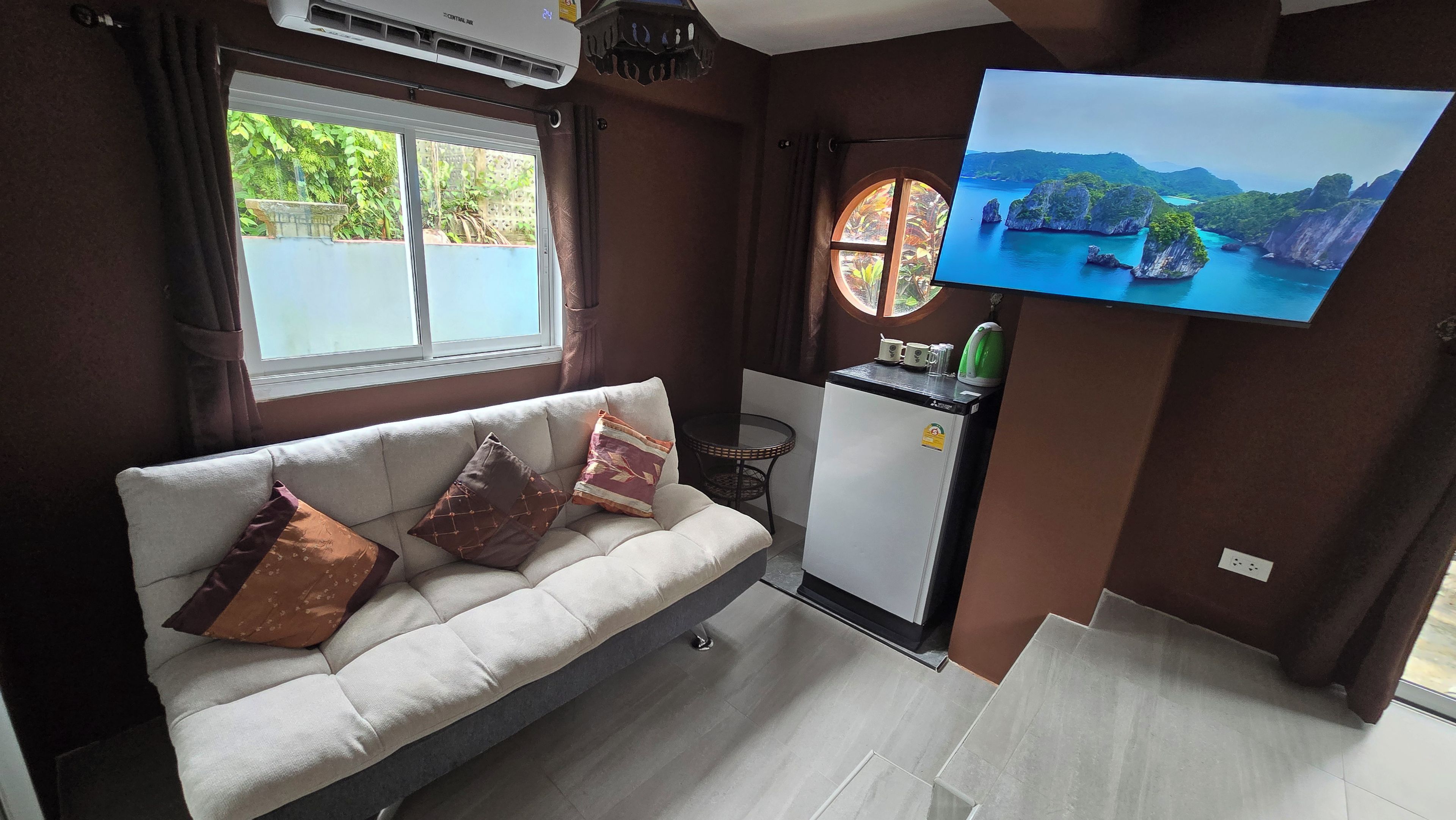 A compact living space with a sofa, a small refrigerator, a television, and a window with a view of greenery.