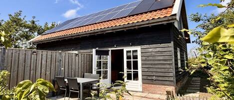 A wooden house with a solar panel roof, a patio with a table and chairs, and a wooden fence.