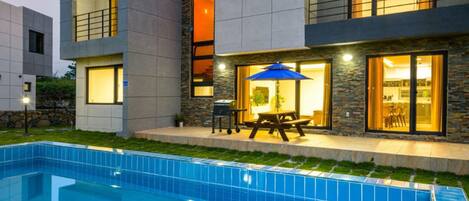 A modern two-story house with a swimming pool, outdoor seating, and large windows.