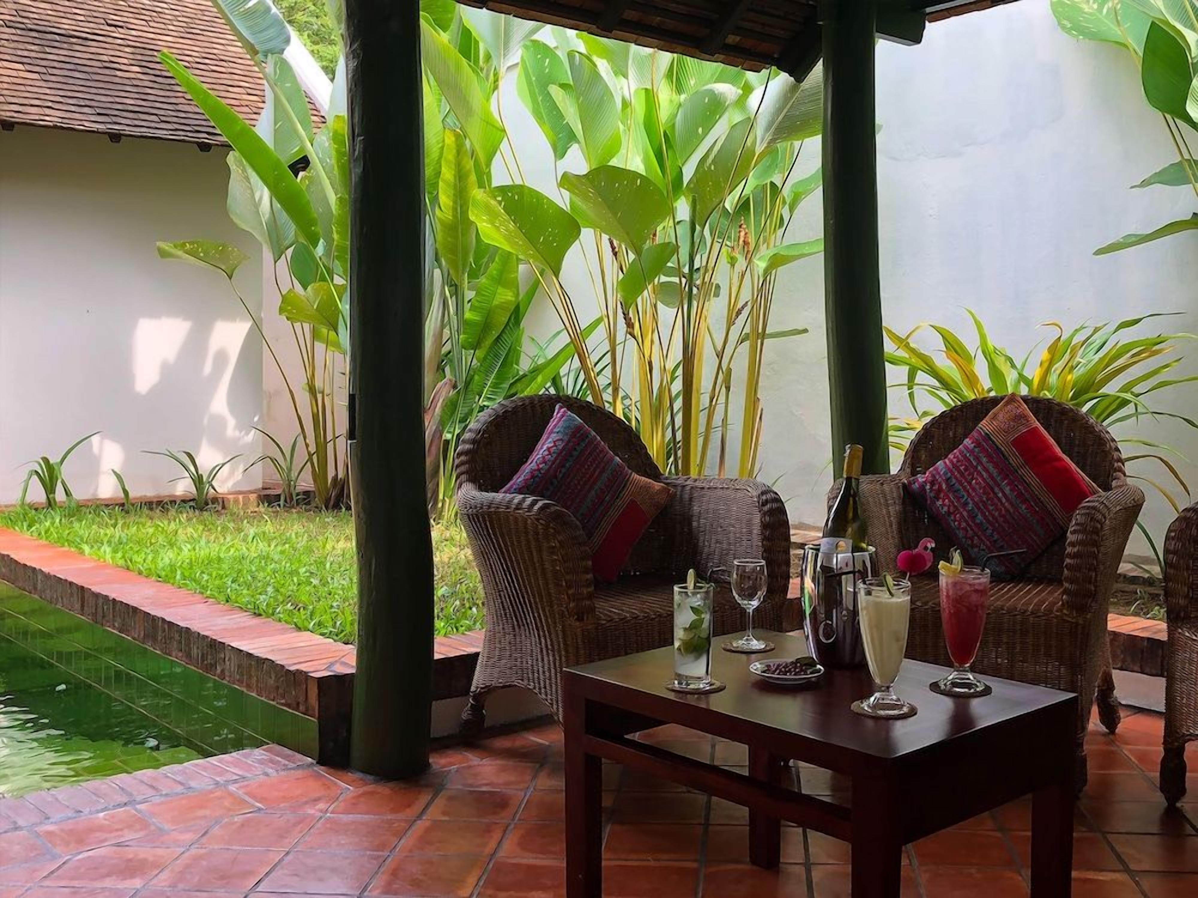 Un espacio de estar al aire libre cubierto con mobiliario de mimbre y una mesa dispuesta para dos personas, rodeado de exuberante follaje y una pequeña fuente de agua.