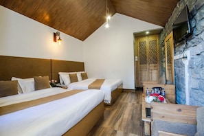 Quarto de hotel com duas camas, cabeceira de madeira, parede de pedra, televisão e uma pequena mesa com garrafas e flores.