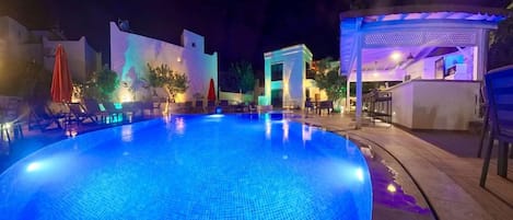 A nighttime swimming pool area with lounge chairs and a covered seating area.