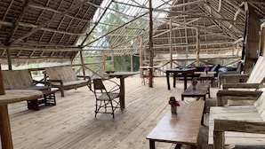 An open-air wooden deck with seating arrangements and tables, surrounded by a thatched roof structure. - Jambo Beach Bungalows - Hostel (Paje)