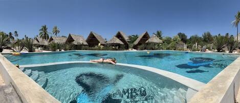 A swimming pool with a person in the water, surrounded by thatched-roof structures and palm trees.