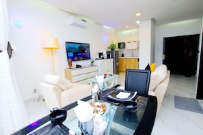 Una sala de estar moderna con una mesa de comedor de vidrio, sofás, un televisor de pantalla plana y una cocina al fondo.