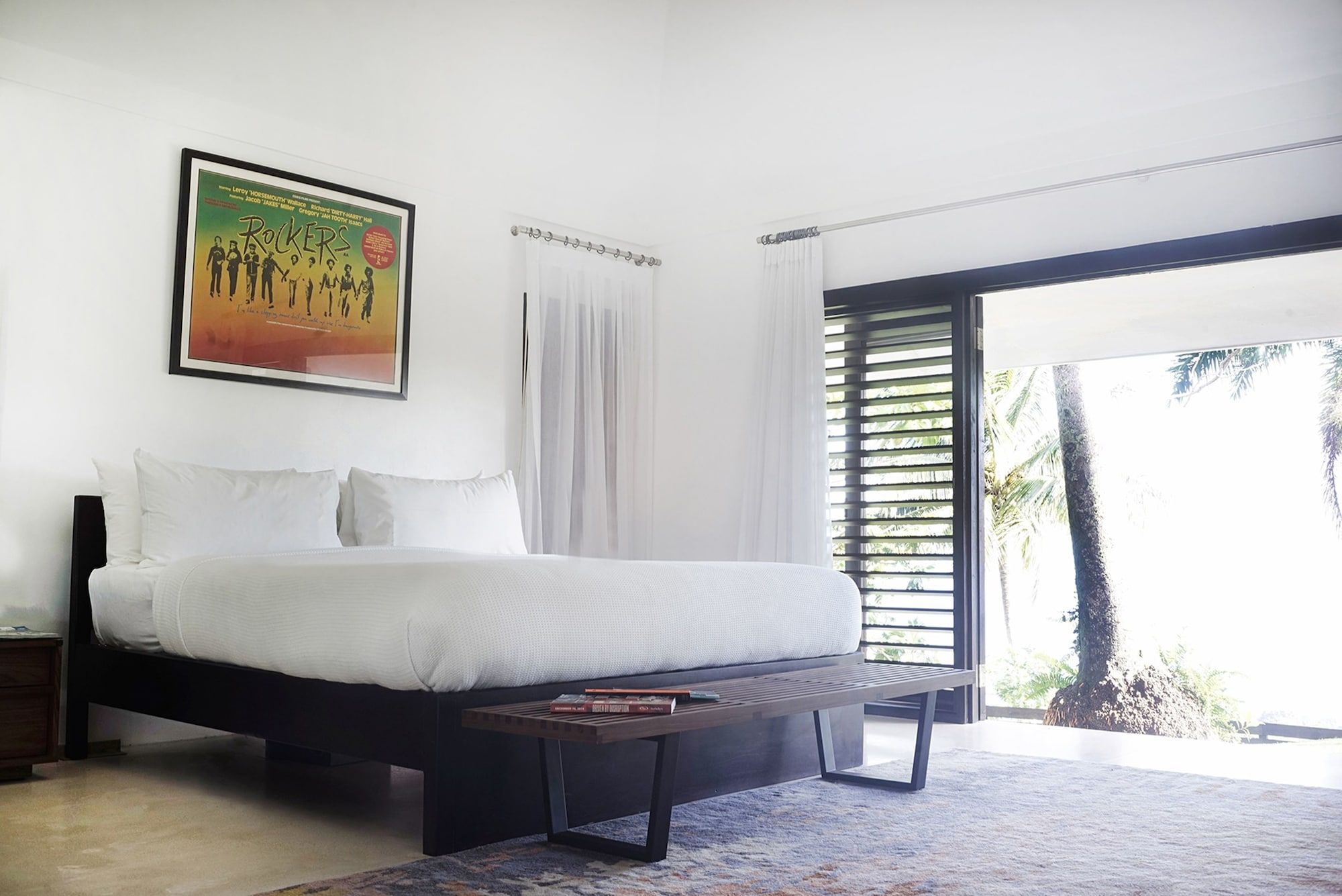 A bedroom with a bed, a bedside table, a framed poster, and a view of palm trees through a window.
