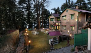 A two-story building with a brick exterior, multiple windows with green trim, and a well-lit outdoor seating area with tables and chairs. - Brij Villa, Dalhousie (Dalhousie)