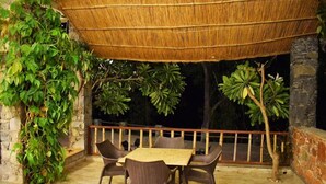 An outdoor seating area with a table and chairs, surrounded by greenery and a thatched roof.