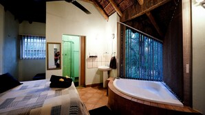 Una habitación con cama, puerta verde, baño con lavamanos y bañera, y ventana con persianas verticales.