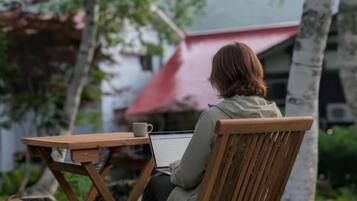 A person sitting at an outdoor wooden table with a laptop, surrounded by greenery and trees.