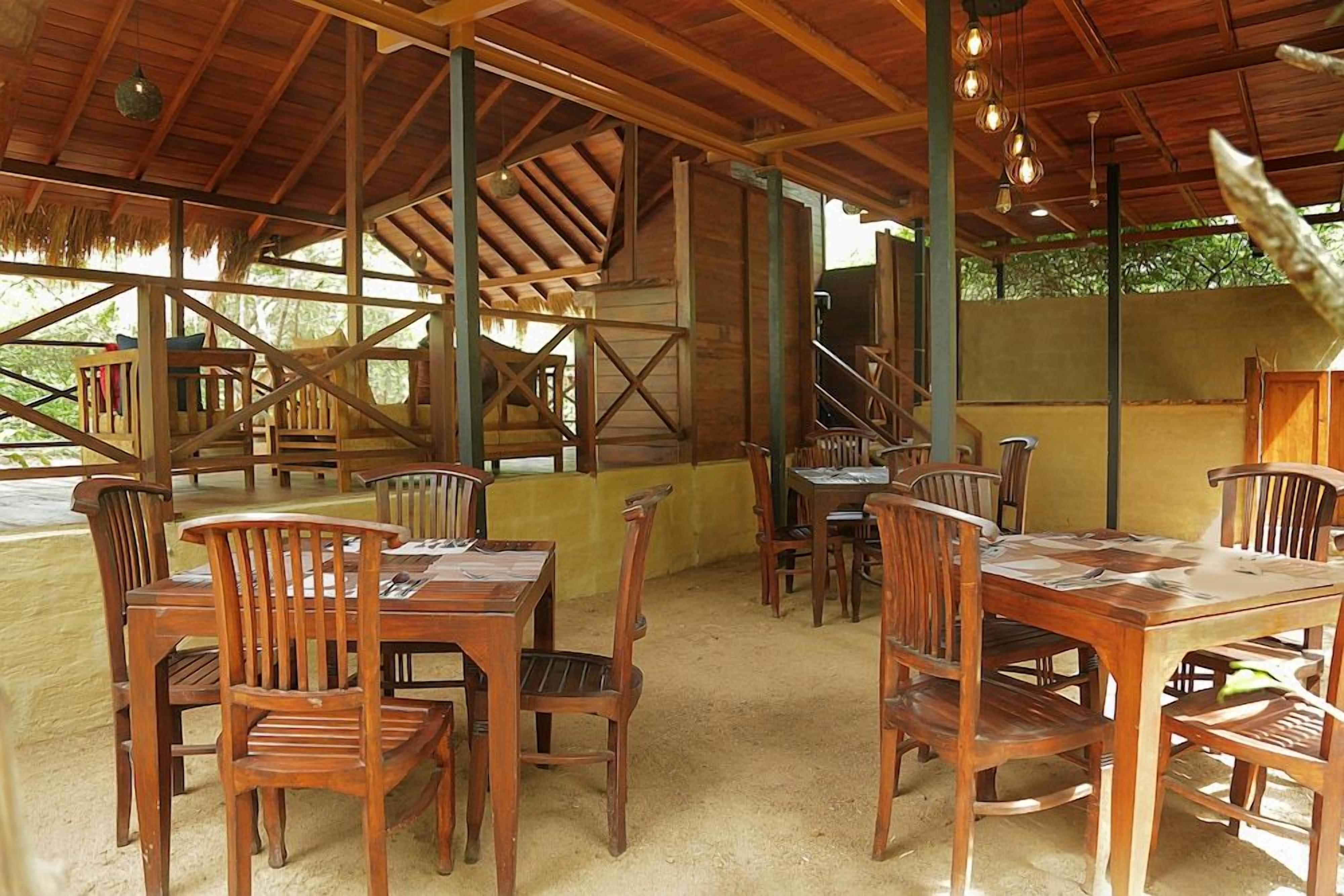 An outdoor wooden dining area with tables and chairs, surrounded by a thatched roof structure.